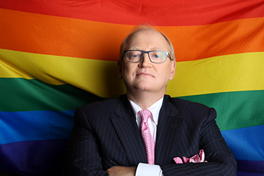 Douglas Elliott stands with arms folded in front of a rainbow flag