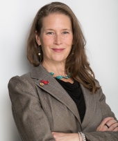 White woman with blue eyes, red lips, and shoulder-length light brown hair wearing grey blazer, black shirt, turquoise beaded necklace, smiling and standing with arms folded